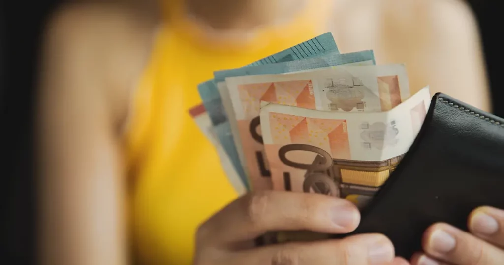 Persona ar maku un izplestām eiro banknotēm rokās, ģērbusies dzeltenā topā.
