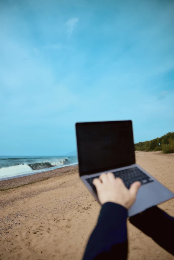 A man holds a laptop in his hand, with the sea, sand and blue sky in the background