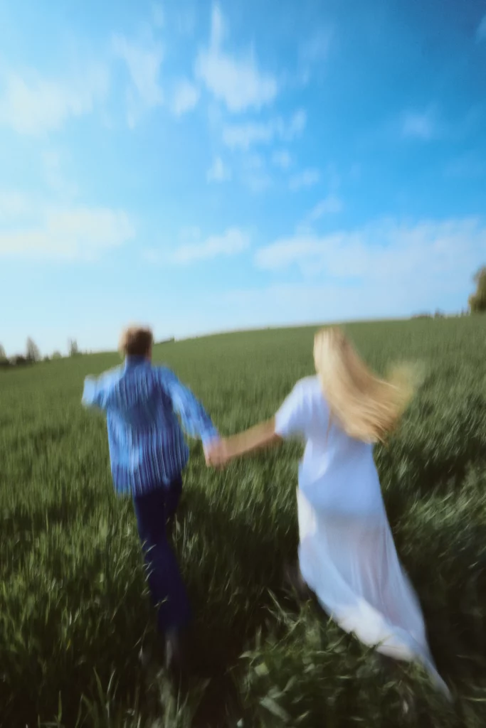A man and a woman holding hands and running in a green field of crops, with a blue sky in the background