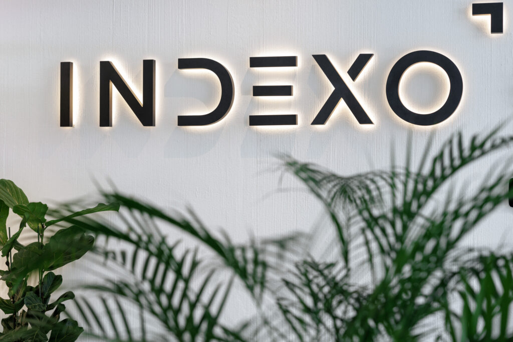 Illuminated INDEXO logo sign on a white wall, green plants in the foreground