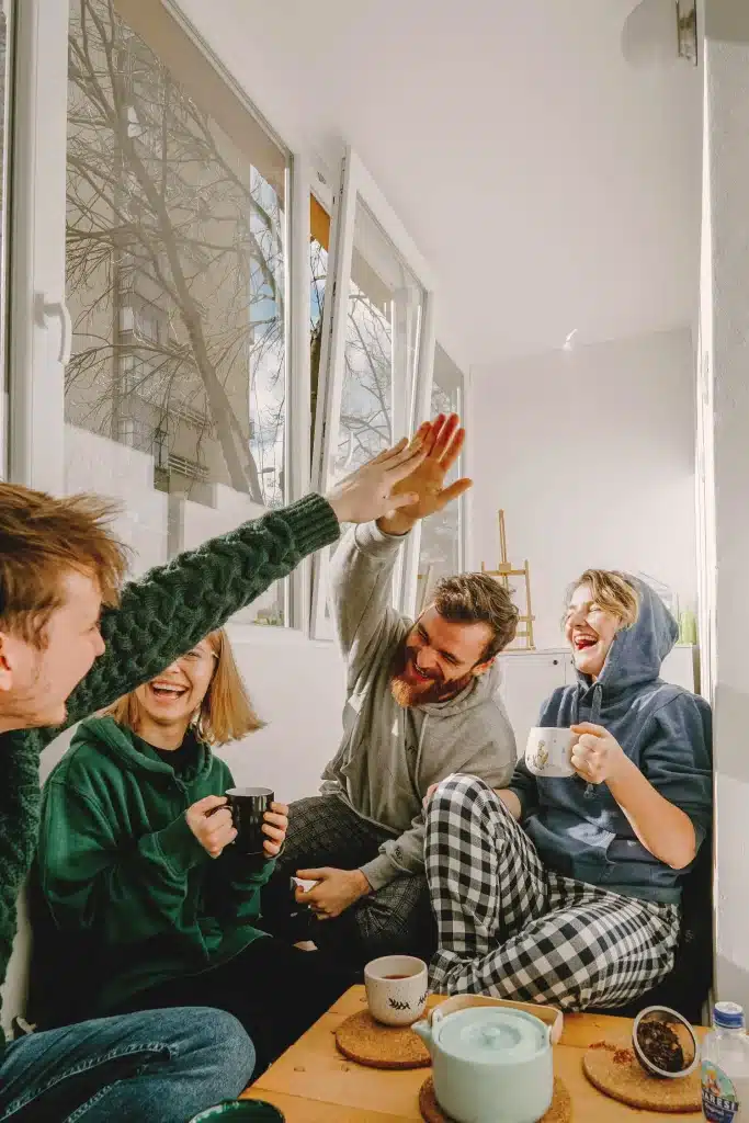 Four friends sitting by a window, laughing, drinking tea, and giving each other high fives.