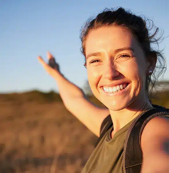 Smiling woman taking a selfie outdoors with her arm raised, standing in a sunny, grassy field.