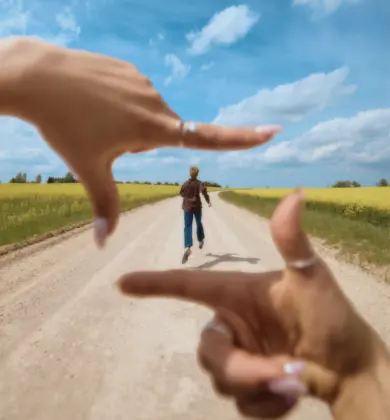 A man runs along a country road, framed between two arms in the foreground, a flowering field and blue sky in the background